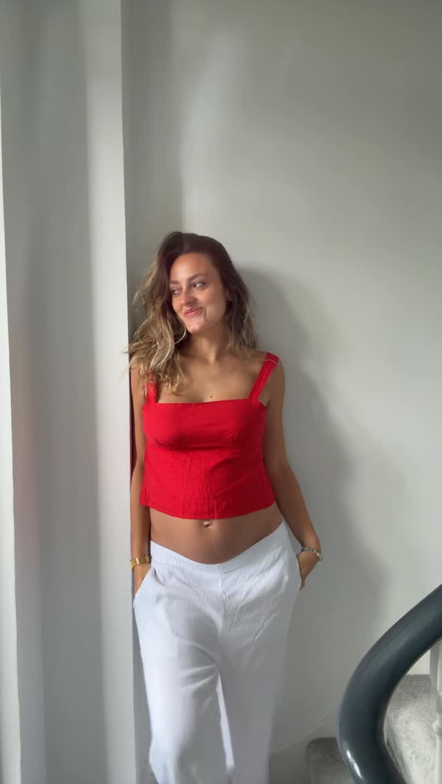Sara in a red corset top and white linen trousers, leaning against a white wall.